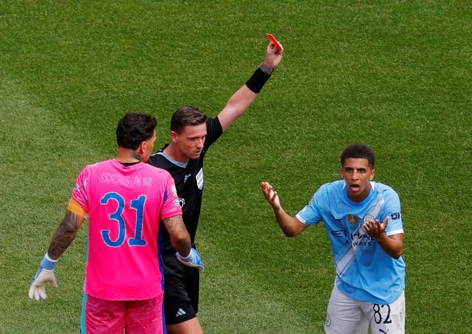 A close-up image of Rico Lewis receiving a red card from the referee, with a look of shock and disbelief on Lewis's face. Show the referee and the surrounding players.