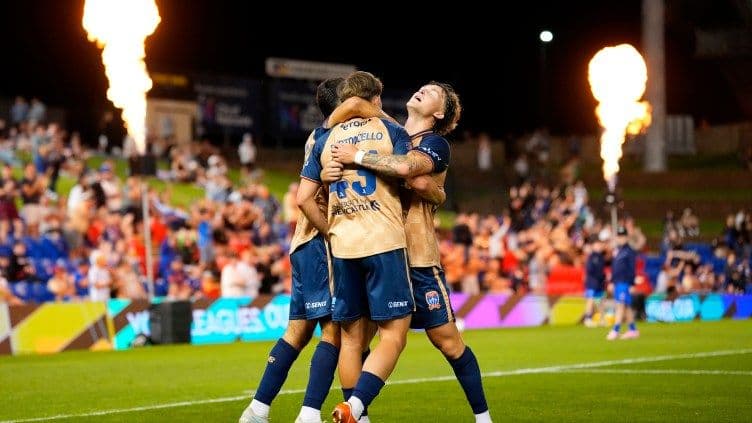 Ba cầu thủ Newcastle Jets ôm nhau ăn mừng bàn thắng trên sân cỏ với pháo sáng phía sau, thể hiện không khí cuồng nhiệt của chuỗi thắng A-League.