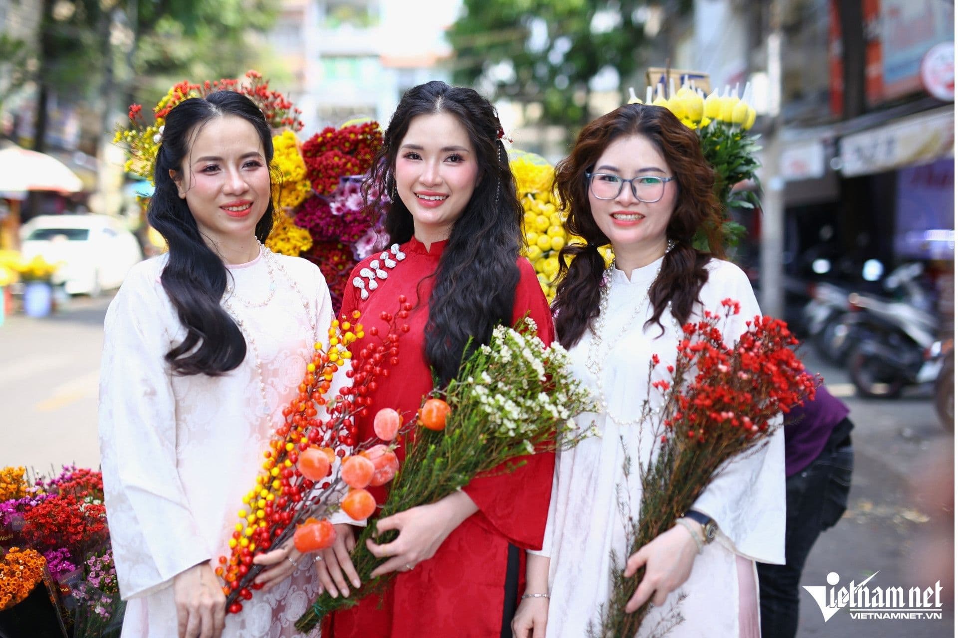 Ba người phụ nữ Việt Nam mặc áo dài truyền thống, tươi cười bên những bó hoa và trái cây rực rỡ, tượng trưng cho không khí Lập Xuân và sự khởi đầu may mắn.