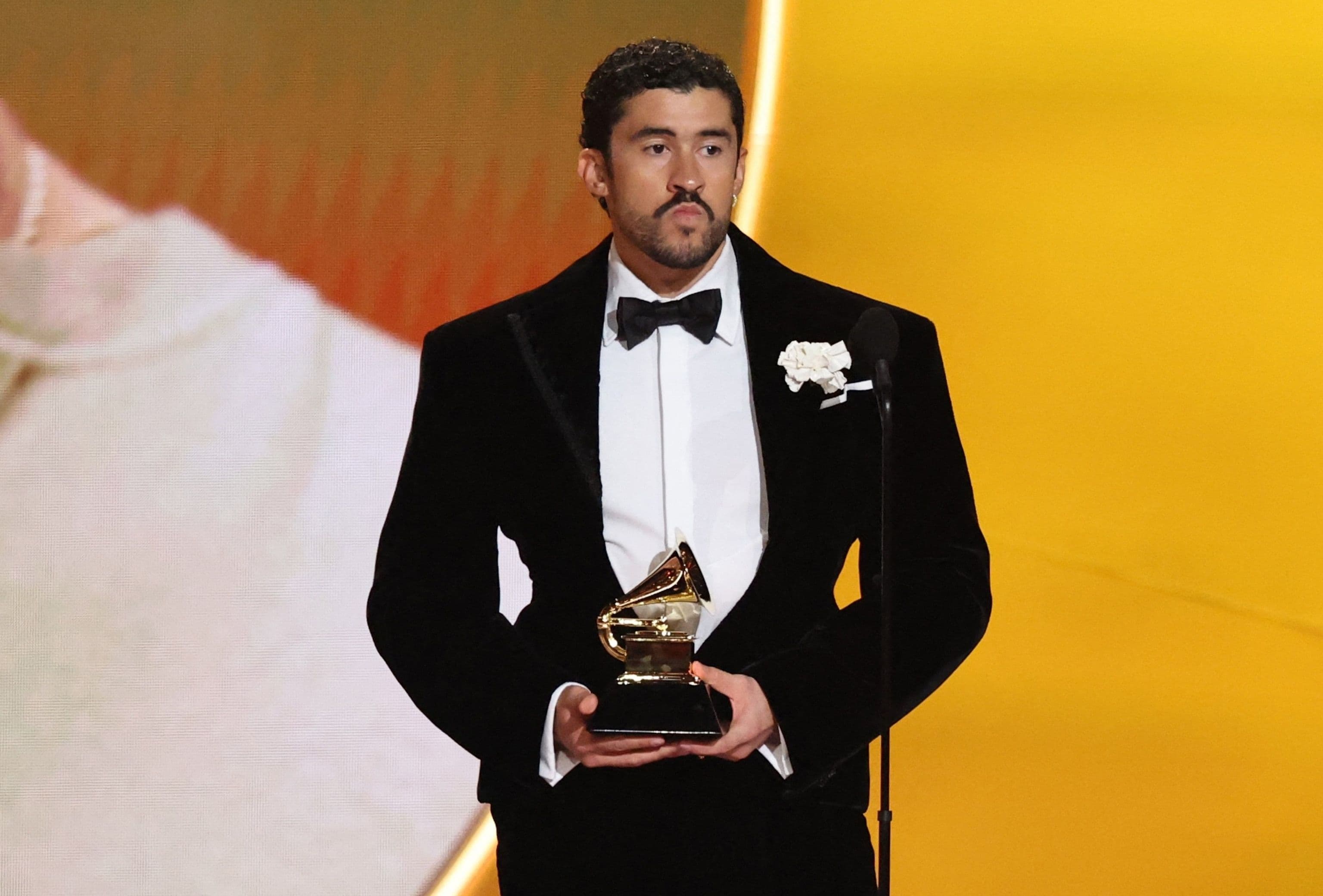 Bad Bunny trong bộ vest đen, cầm giải Grammy, đứng trên sân khấu với phông nền màu vàng.