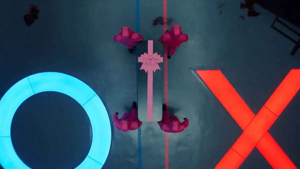 Bốn lính canh áo hồng mang hộp quà giữa sàn đấu với biểu tượng O và X trong Trò Chơi Con Mực.
