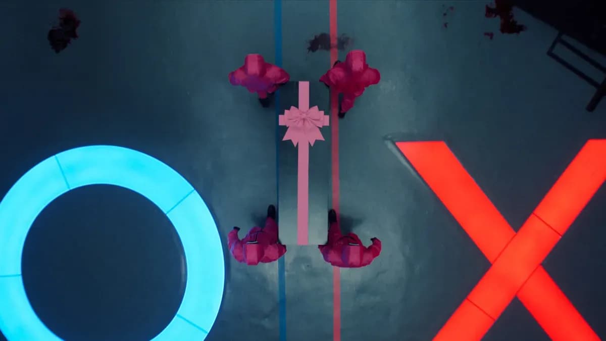 Bốn lính canh áo hồng mang hộp quà trên sân chơi với biểu tượng O và X, tượng trưng cho các trò chơi chết chóc của Trò Chơi Con Mực.