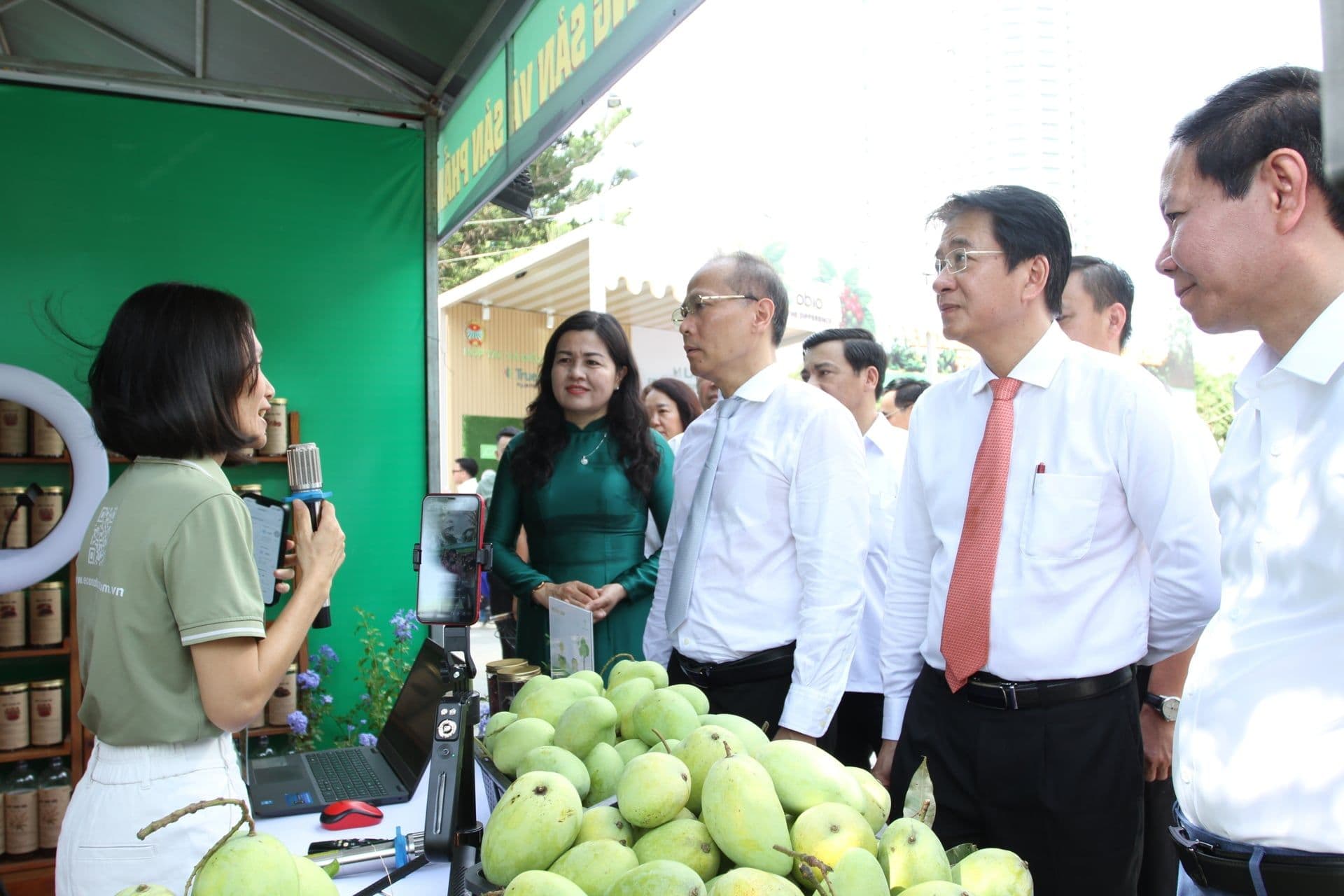 Các quan chức Khánh Hòa tham quan gian hàng trưng bày sản phẩm OCOP, với một phụ nữ đang giới thiệu xoài tươi, nhấn mạnh bản sắc và sự đa dạng của thương hiệu OCOP.