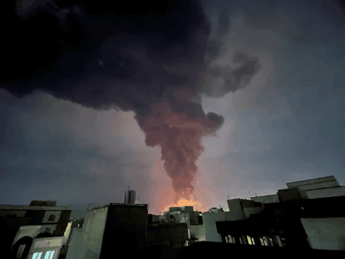 Cột khói lớn bốc lên từ một khu vực đô thị, tượng trưng cho xung đột Trung Đông và tác động đến nguồn cung dầu mỏ.