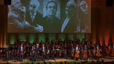 Dàn nhạc giao hưởng Real Filharmonía de Galicia biểu diễn trên sân khấu, phía sau là màn hình lớn chiếu chân dung các nhạc trưởng và nhà soạn nhạc.
