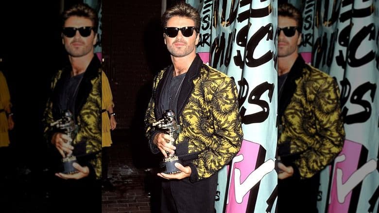 George Michael đeo kính râm, mặc áo khoác vàng họa tiết đen, cầm giải thưởng Vanguard tại VMA 1989, tượng trưng cho di sản của các nghệ sĩ tiên phong trong việc định hình văn hóa thị giác.