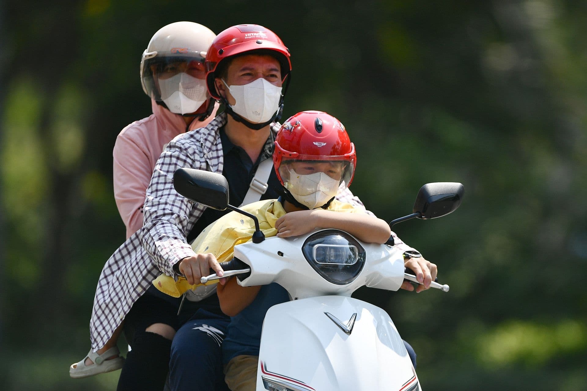 Gia đình ba người đi xe máy dưới trời nắng, đeo khẩu trang và mũ bảo hiểm, thể hiện sự chủ động ứng phó với thời tiết khắc nghiệt.