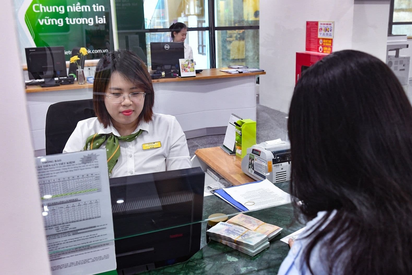 Giao dịch viên ngân hàng Việt Nam đang phục vụ khách hàng, với tiền mặt trên quầy, thể hiện hoạt động ngân hàng và huy động vốn.