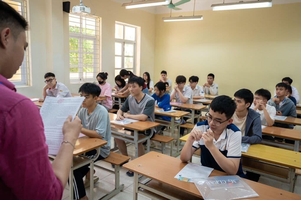 Giám thị đứng trong phòng thi, giám sát các thí sinh đang làm bài, tượng trưng cho niềm tin vào sự công bằng trong giáo dục.