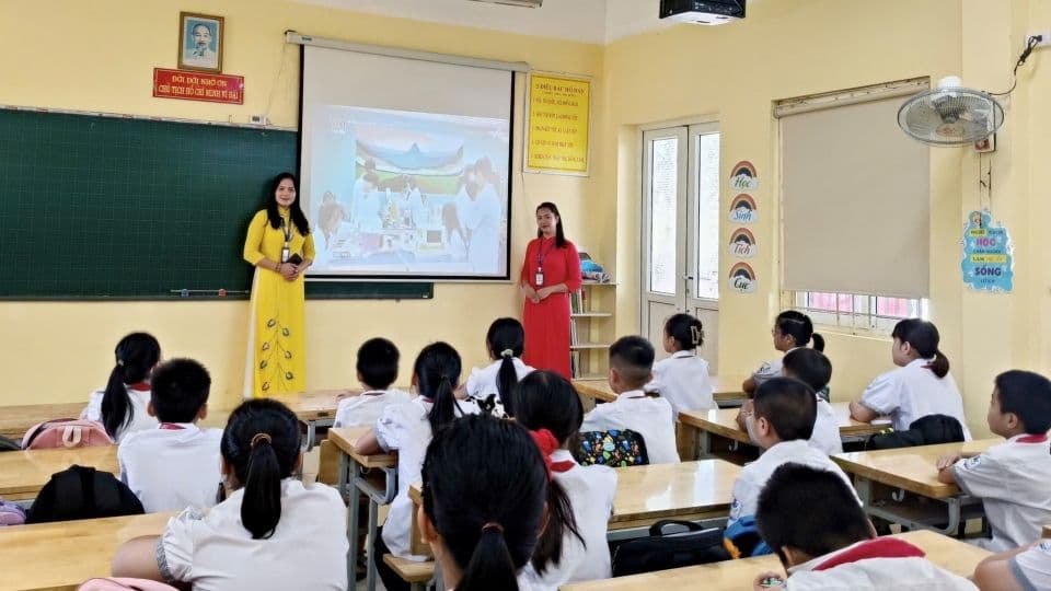 Hai cô giáo đang hướng dẫn học sinh tiểu học trong lớp, nhấn mạnh vai trò của giáo viên và học sinh trong quá trình sáp nhập trường học.