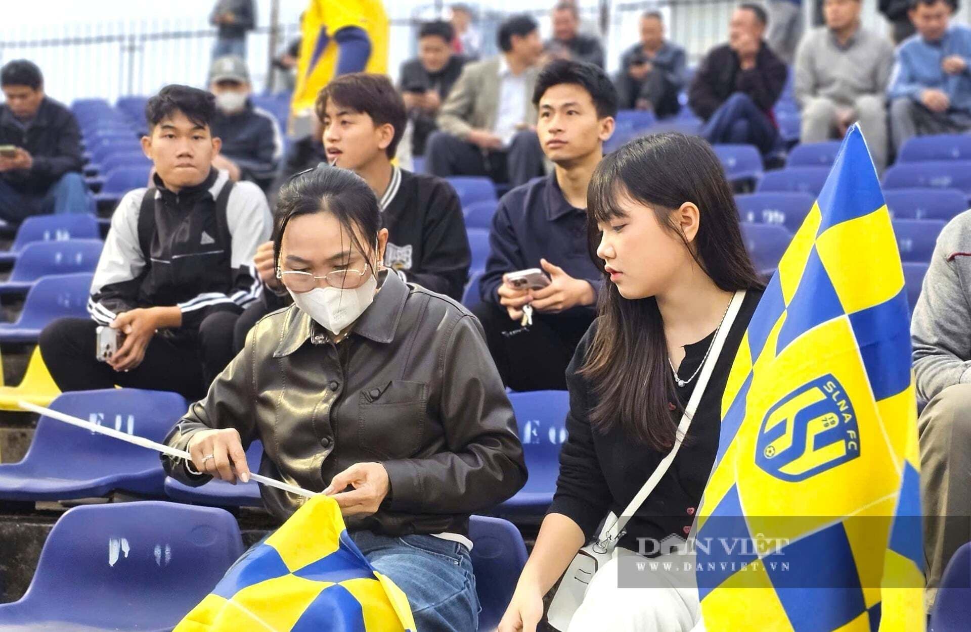 Hai nữ cổ động viên Sông Lam Nghệ An trên khán đài, một người cầm cờ đội bóng, thể hiện sự ủng hộ cuồng nhiệt.