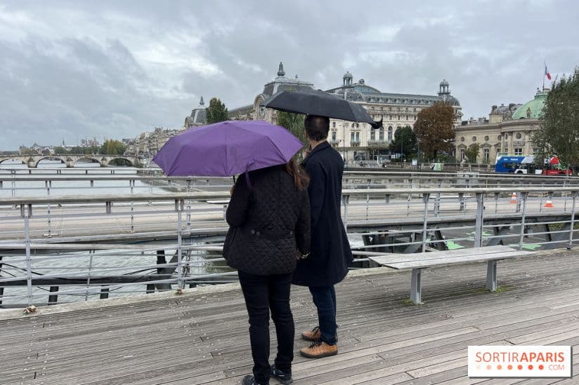 Hai người che ô đi dạo bên sông Seine ở Paris dưới bầu trời nhiều mây, với bảo tàng Orsay phía sau, minh họa sự thay đổi thời tiết thất thường.