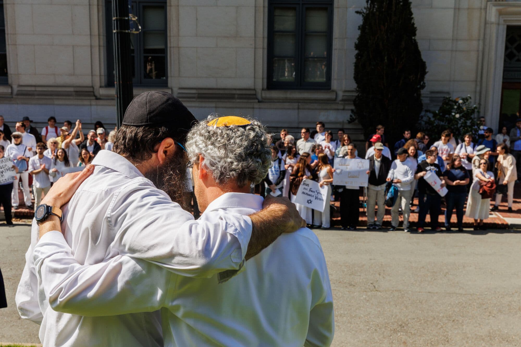 Hai người đàn ông Do Thái ôm nhau trong một cuộc biểu tình, phía sau là đám đông với các biểu ngữ, thể hiện sự đoàn kết trong bối cảnh làn sóng chống thù ghét.
