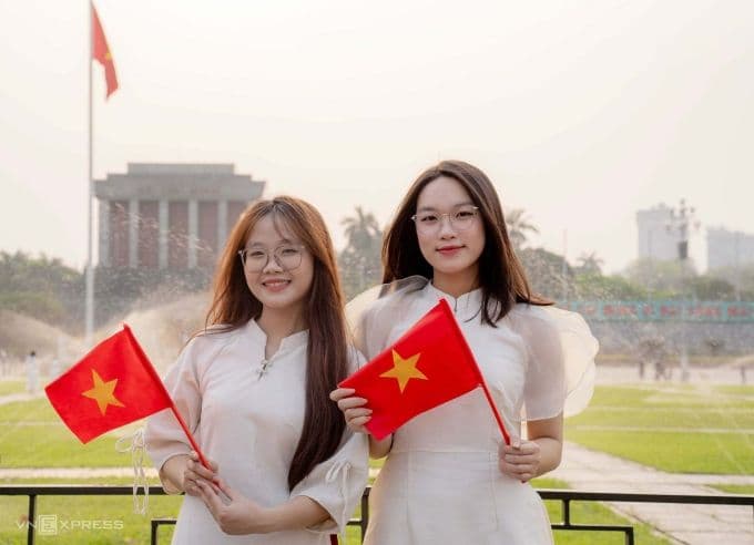 Hai thiếu nữ Việt Nam mặc áo dài, cầm cờ Tổ quốc tại quảng trường Ba Đình, Hà Nội, tượng trưng cho ký ức lịch sử và nhịp sống hiện đại.