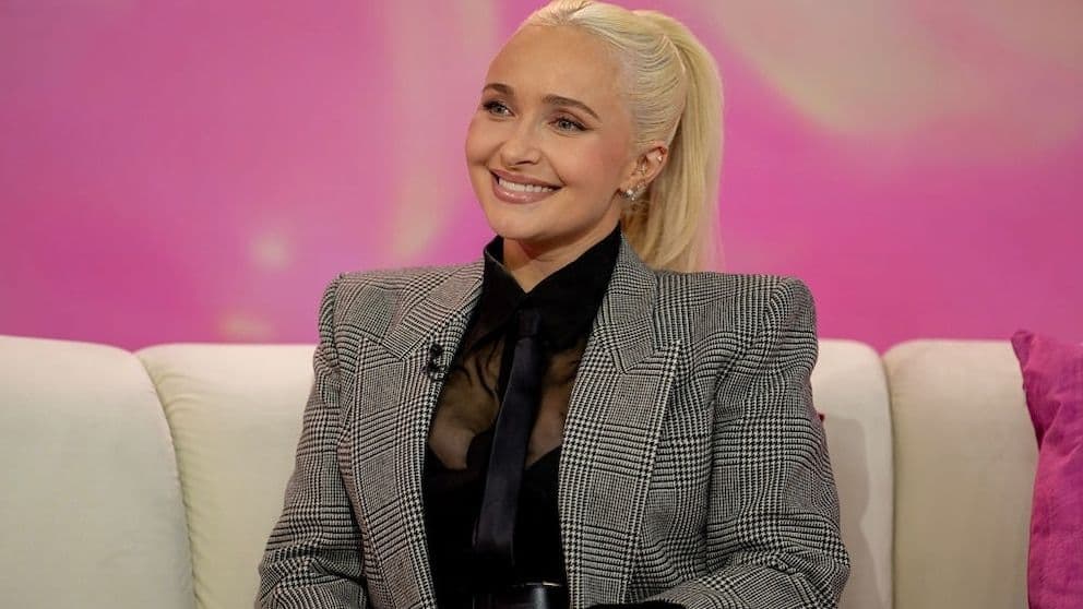 Hayden Panettiere mỉm cười, tóc vàng buộc cao, mặc áo blazer kẻ caro, đại diện cho cuộc đời đầy ánh hào quang và thử thách.