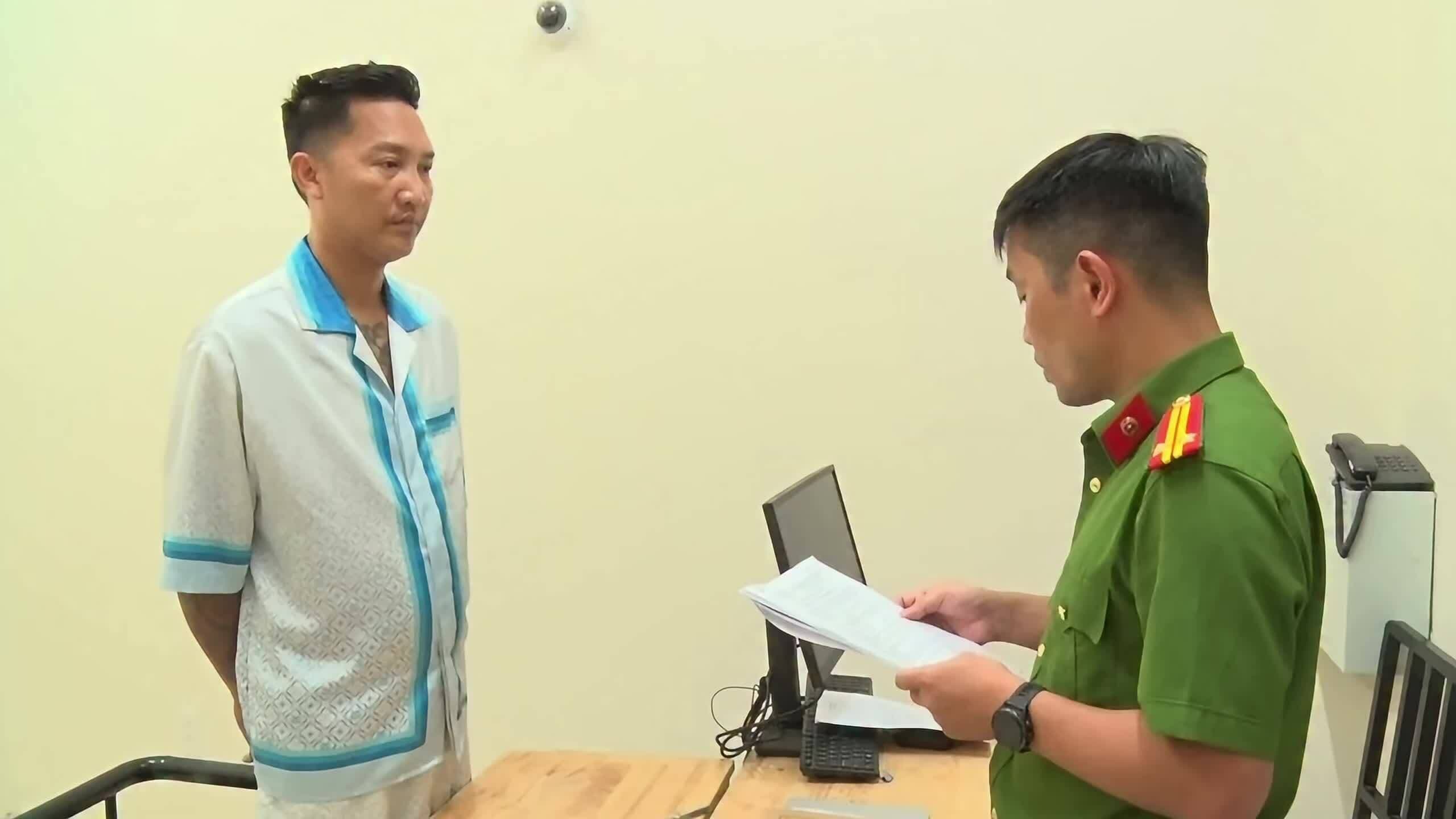 Huấn Hoa Hồng đứng trước cán bộ công an đang xem xét hồ sơ, minh họa quá trình bị cáo buộc hình sự.