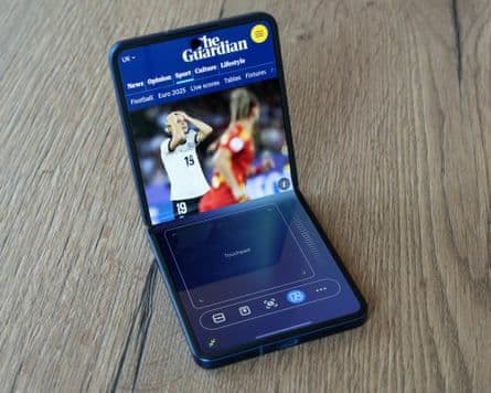 Điện thoại Samsung Galaxy Z Flip7 đang hiển thị trang web The Guardian ở chế độ Flex Mode, với nội dung trên nửa trên và điều khiển cảm ứng ở nửa dưới.