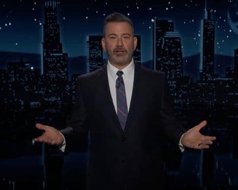 Jimmy Kimmel trên sân khấu chương trình đêm khuya, tay mở rộng, với phông nền thành phố.