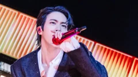 Jin, một ngôi sao K-pop, biểu diễn trên sân khấu với micro đỏ, tượng trưng cho kết nối cảm xúc giữa nghệ sĩ và khán giả trong các buổi hòa nhạc trực tuyến.
