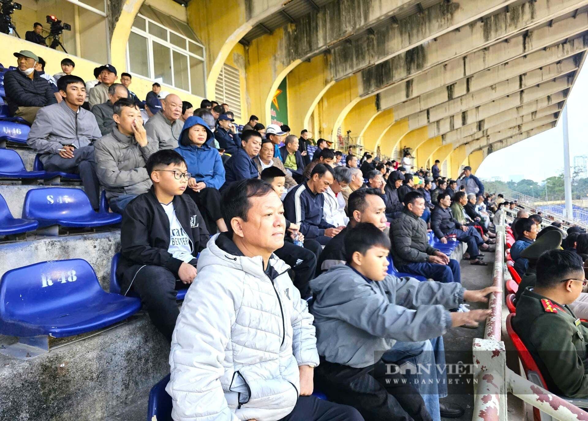 Khán giả ngồi trên khán đài sân vận động, theo dõi trận đấu bóng đá, thể hiện không khí sôi động và tình yêu bóng đá.