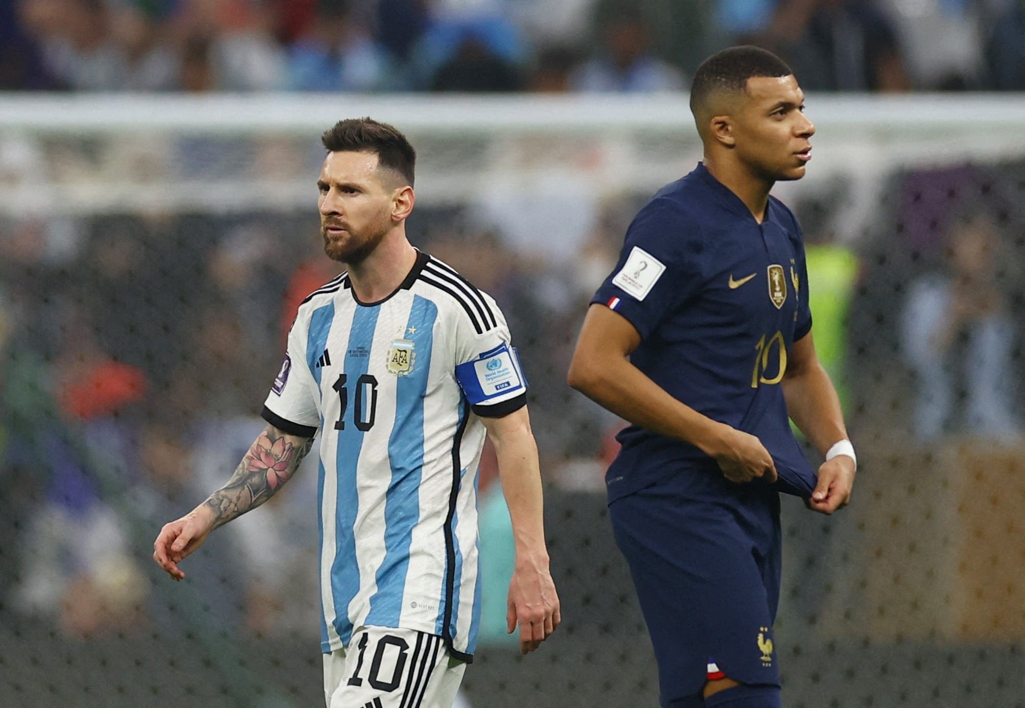 Kylian Mbappé đứng trên sân với vẻ mặt trầm tư khi Lionel Messi ăn mừng bàn thắng trong trận chung kết World Cup 2022, thể hiện khoảnh khắc định hình sự nghiệp.