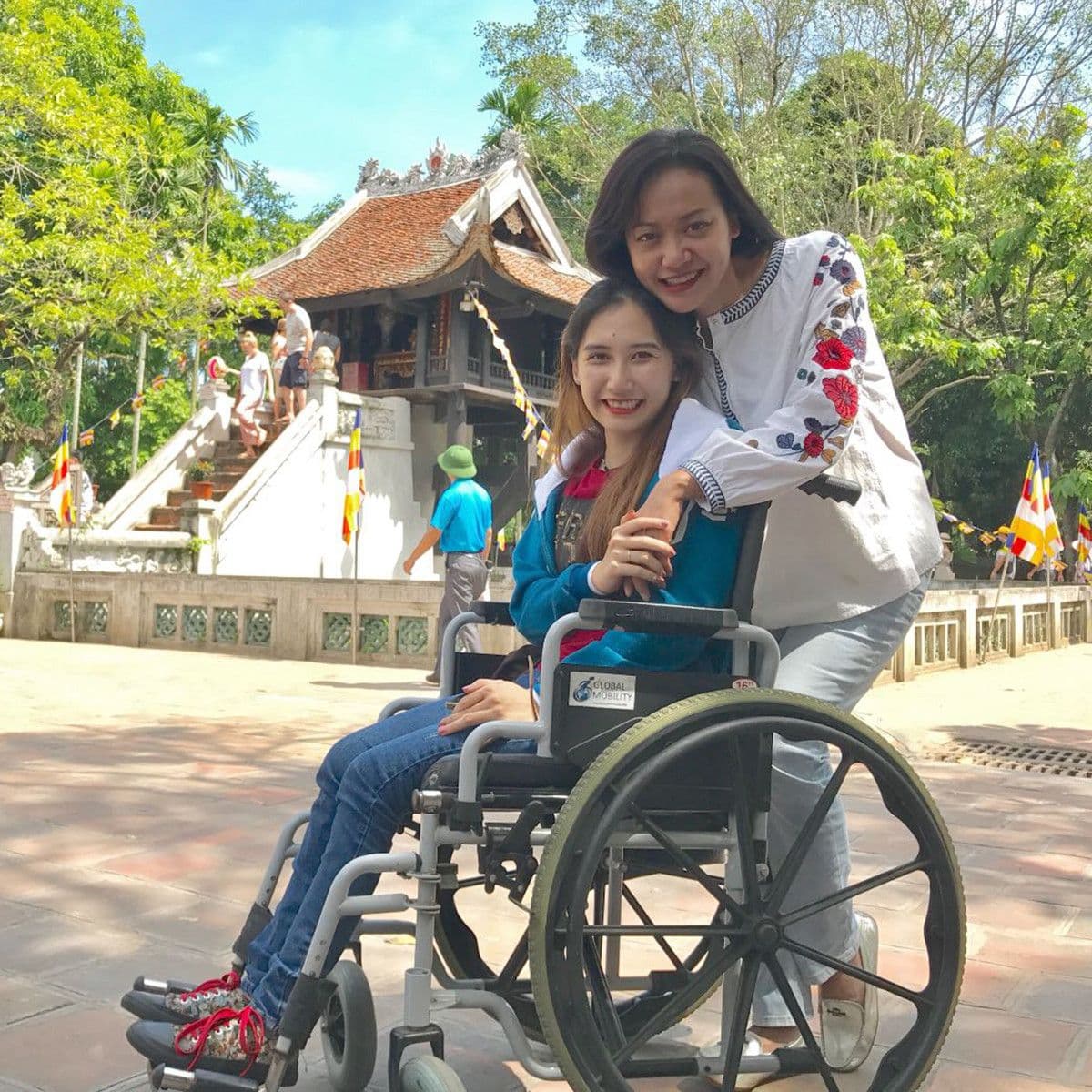 Lê Hiền Hạnh ngồi xe lăn và Hồng Ánh ôm cô từ phía sau, tươi cười trong khung cảnh ngoài trời