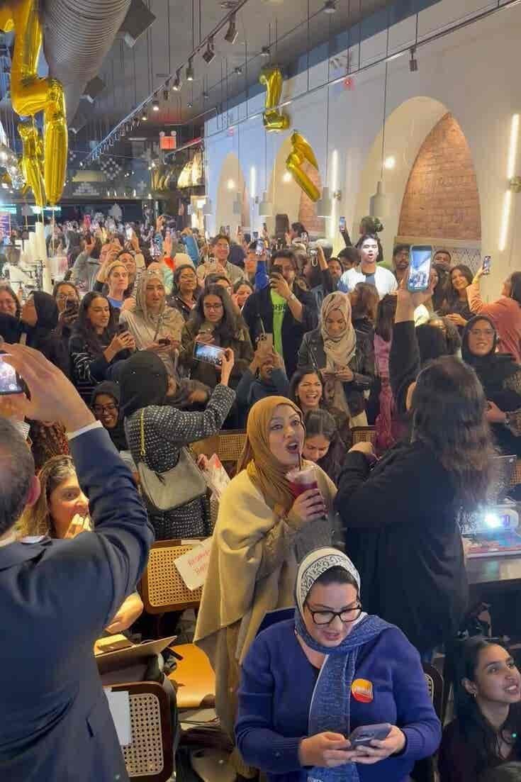 Đám đông đa dạng tại một quán cà phê ở New York đang reo hò và ghi lại khoảnh khắc chiến thắng của Zohran Mamdani bằng điện thoại.