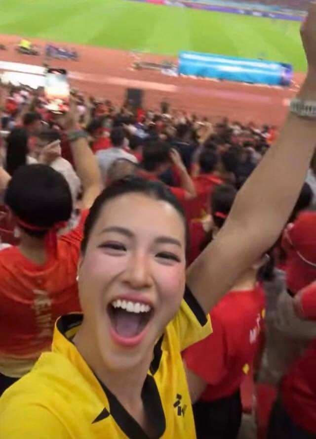 MC Hoàng Oanh mặc áo đấu màu vàng, tươi cười cổ vũ trên khán đài, thể hiện sự tự tin và lạc quan.