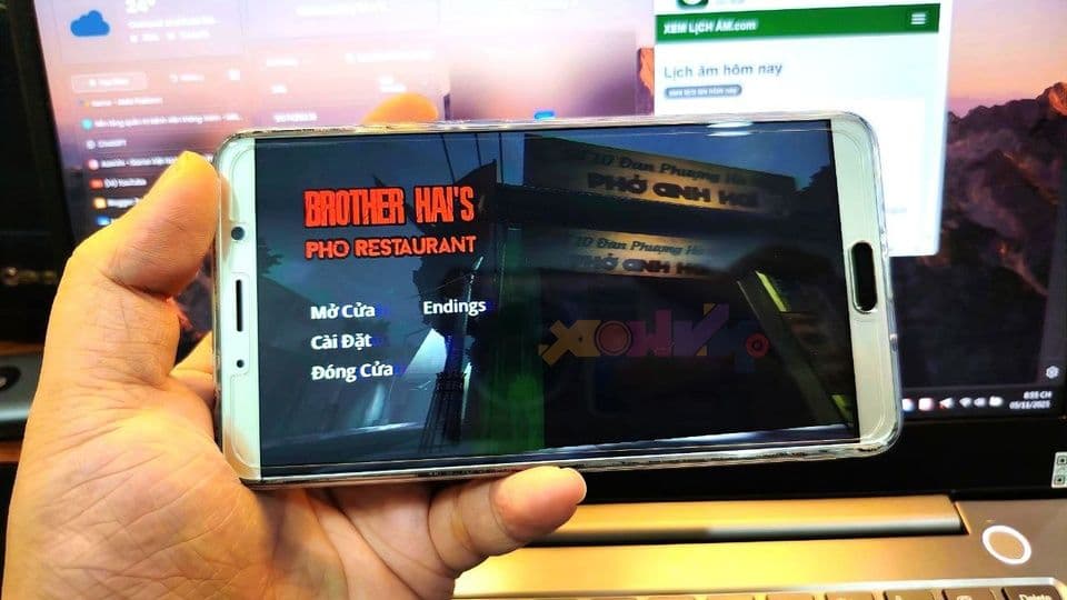 Màn hình điện thoại hiển thị trò chơi 'Tiệm Phở của Anh Hai' trên nền laptop.