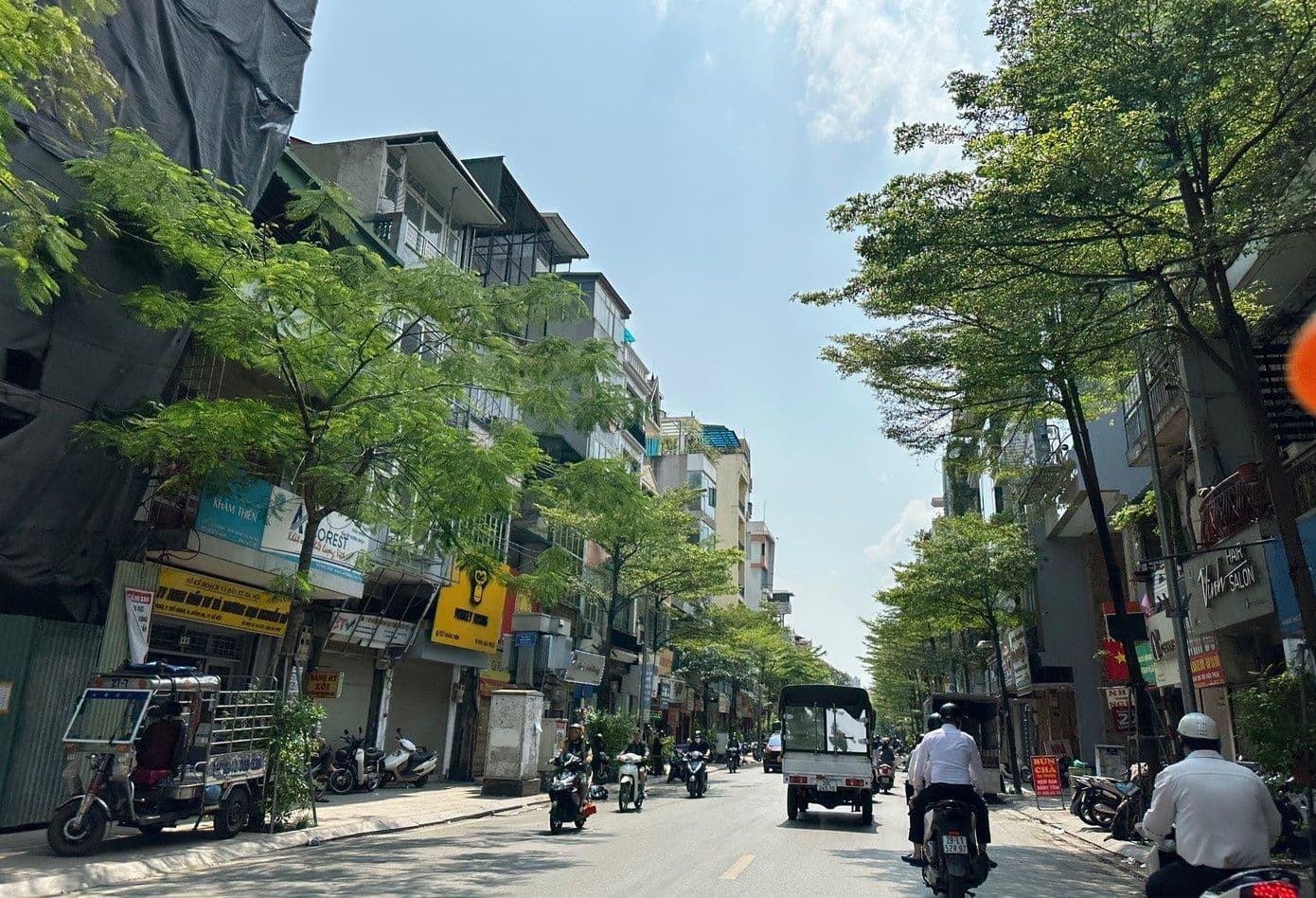 Một con phố Hà Nội với nắng nhẹ, cây xanh và người dân di chuyển, thể hiện thời tiết đẹp.