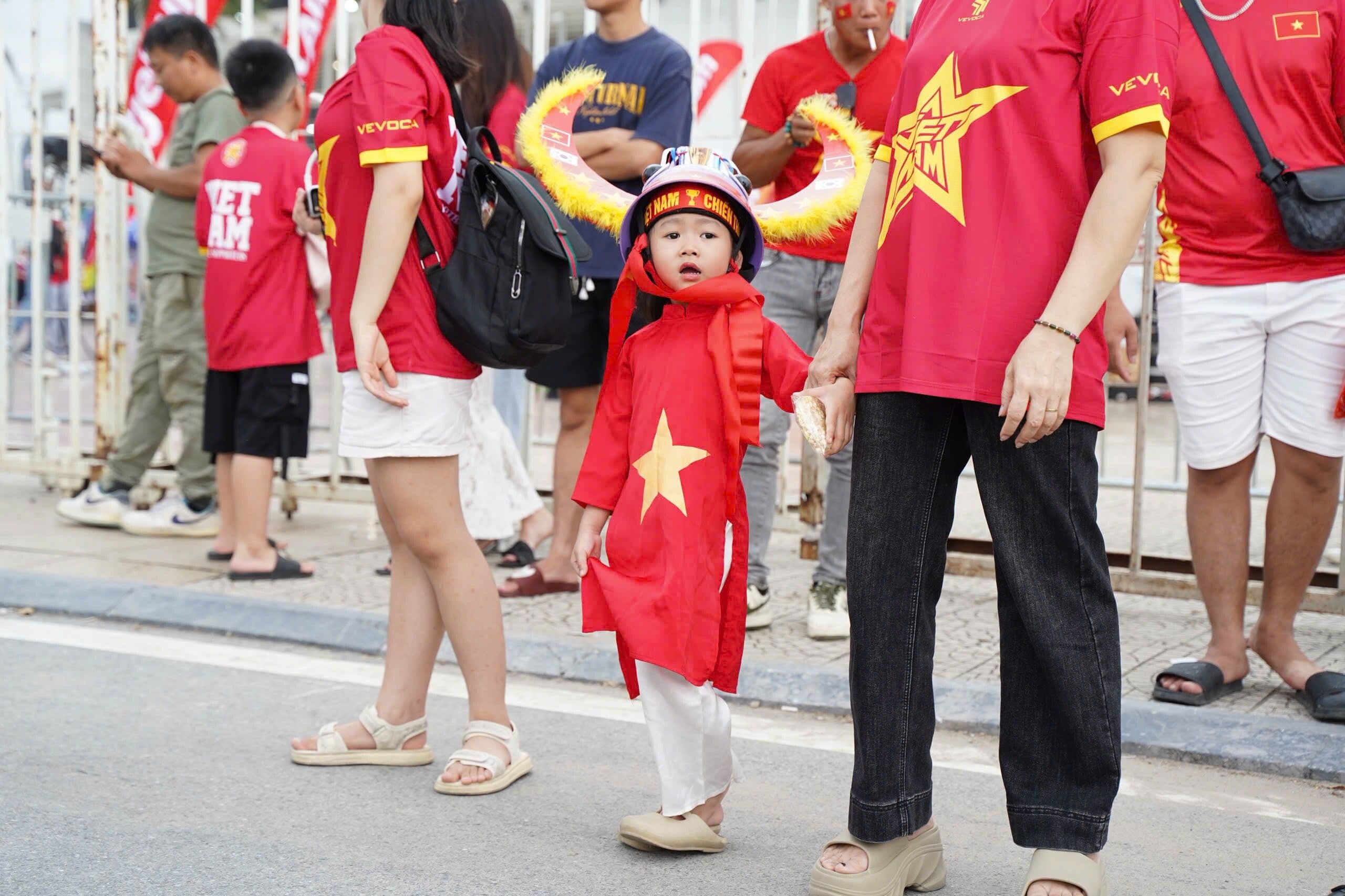 Một em bé mặc áo đội tuyển Việt Nam, đội mũ có cánh, cùng người lớn tham gia cổ vũ bóng đá, thể hiện sự kết nối thế hệ.