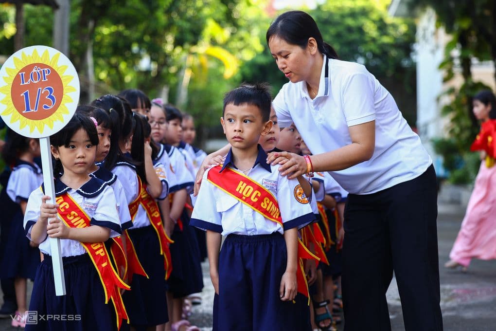 Một giáo viên đang chỉnh trang cho học sinh tiểu học xếp hàng, thể hiện sự quan tâm đến thế hệ tương lai trong bối cảnh tái cấu trúc trường học.