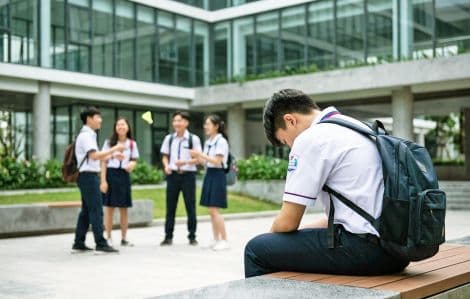 Một học sinh ngồi một mình, buồn bã, trong khi những học sinh khác đang trò chuyện, tượng trưng cho sự cô lập và xói mòn niềm tin do tin giả.