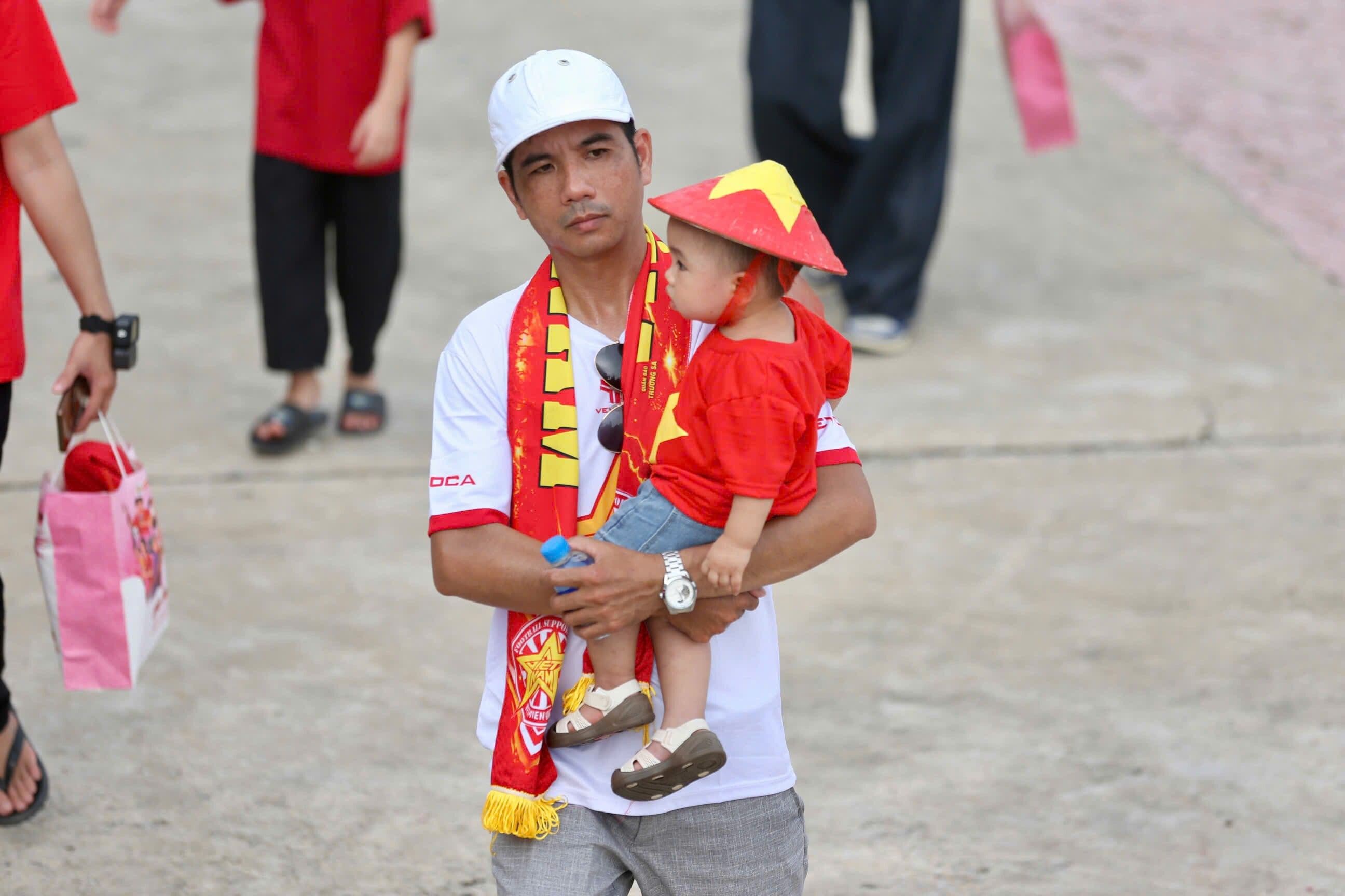 Một người cha bế con nhỏ, cả hai mặc áo và đội nón cờ Việt Nam, thể hiện tình yêu bóng đá qua các thế hệ.