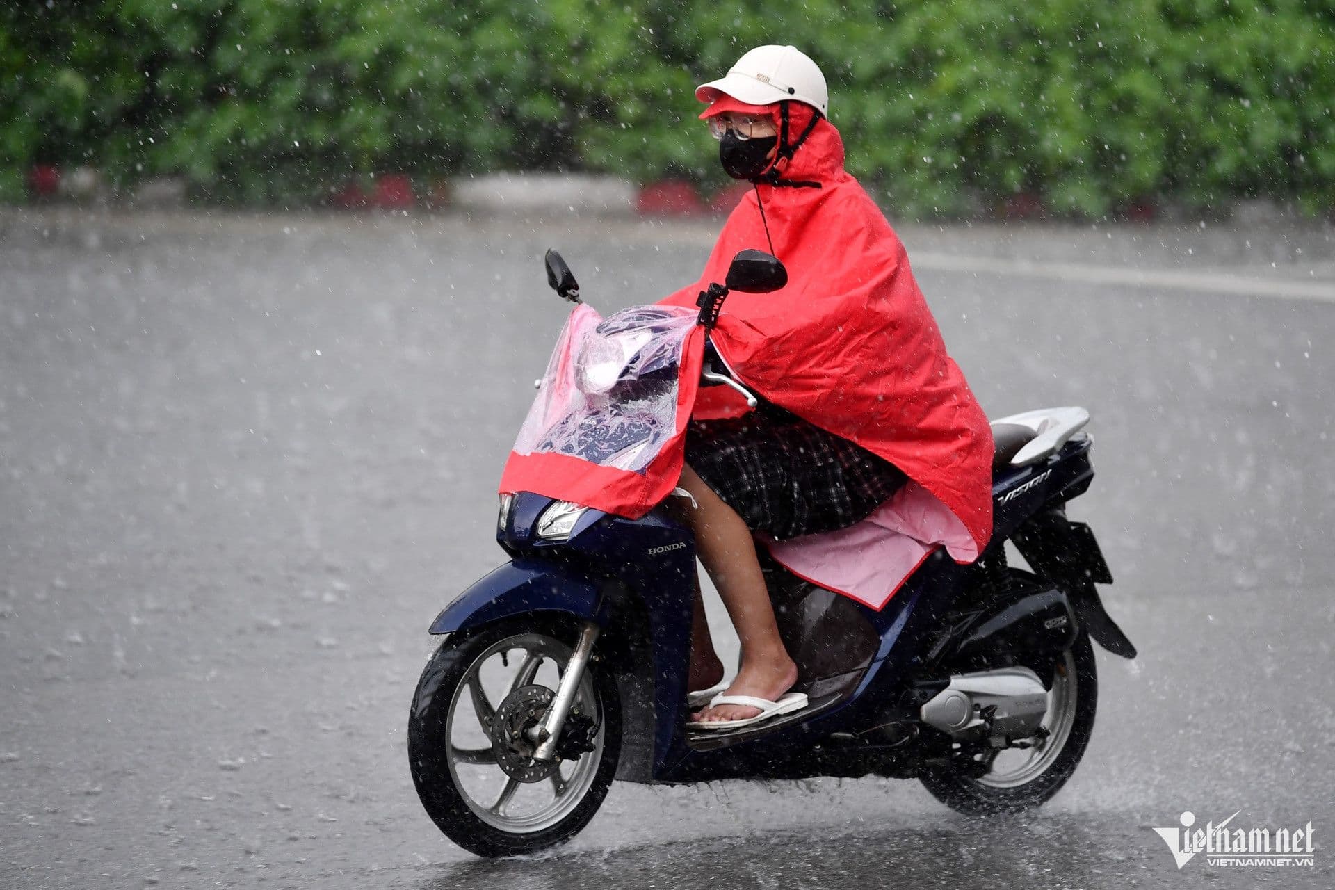 Một người đi xe máy mặc áo mưa màu đỏ trên đường ướt, thể hiện sự thích nghi cá nhân với thời tiết mưa.