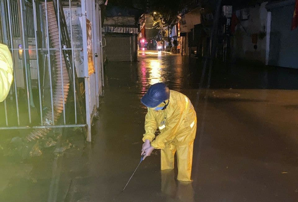 Một người mặc áo mưa đang kiểm tra cống thoát nước trên con đường ngập, minh họa nỗ lực ứng phó với tình hình mưa lũ.