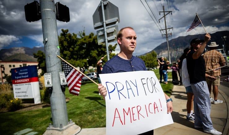 Một người đàn ông cầm biểu ngữ 'PRAY FOR AMERICA!' cùng cờ Mỹ, thể hiện sự đau buồn và phản ứng chính trị sau vụ việc.