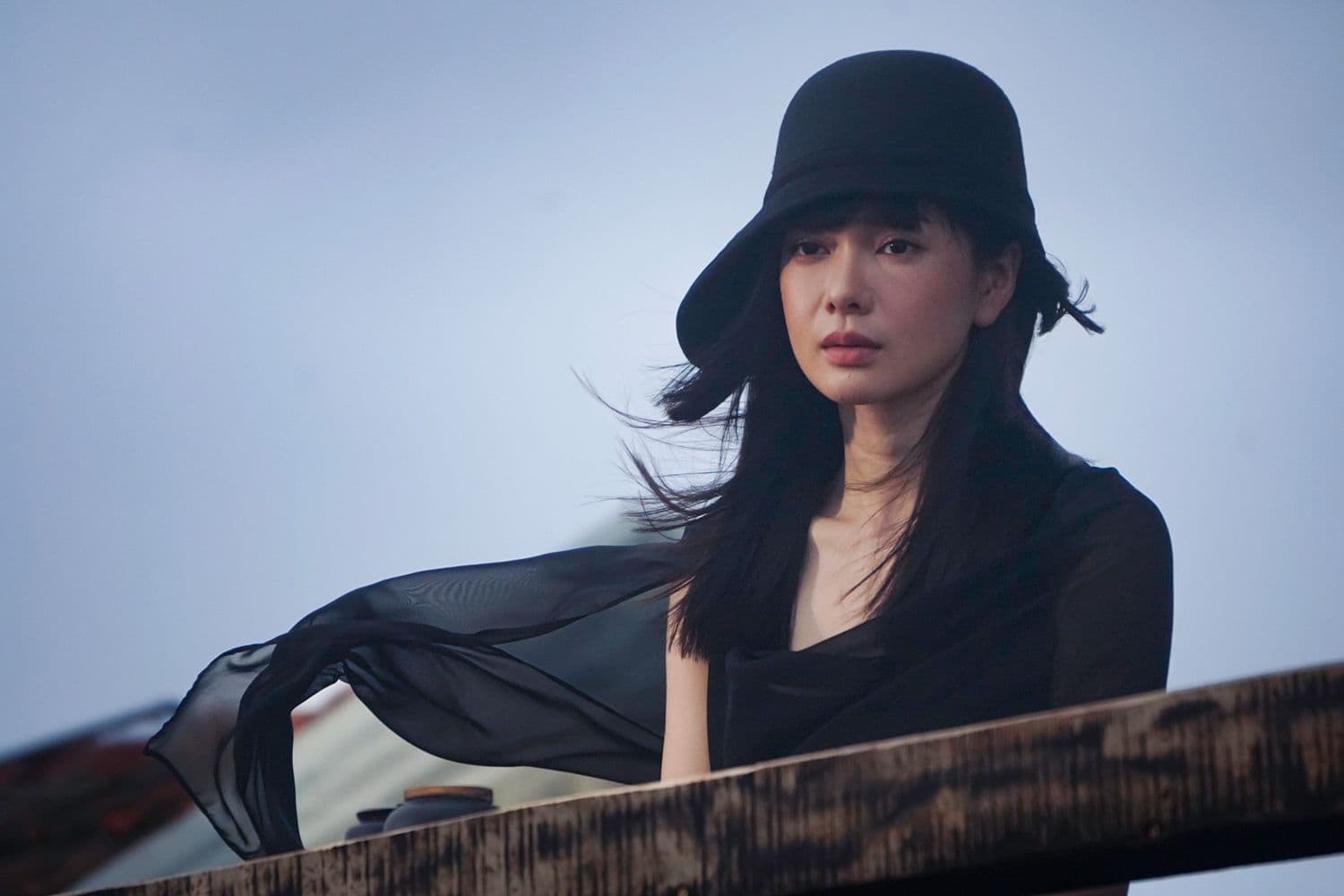 Một người phụ nữ trong trang phục đen và mũ rộng vành, ánh mắt trầm tư, gợi lên sự bí ẩn và nội tâm sâu sắc.
