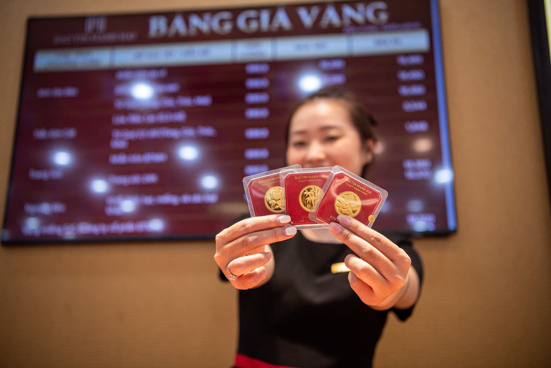 Một nhân viên đang trình bày các sản phẩm vàng trước bảng giá vàng điện tử, thể hiện sự biến động của thị trường vàng hôm nay.