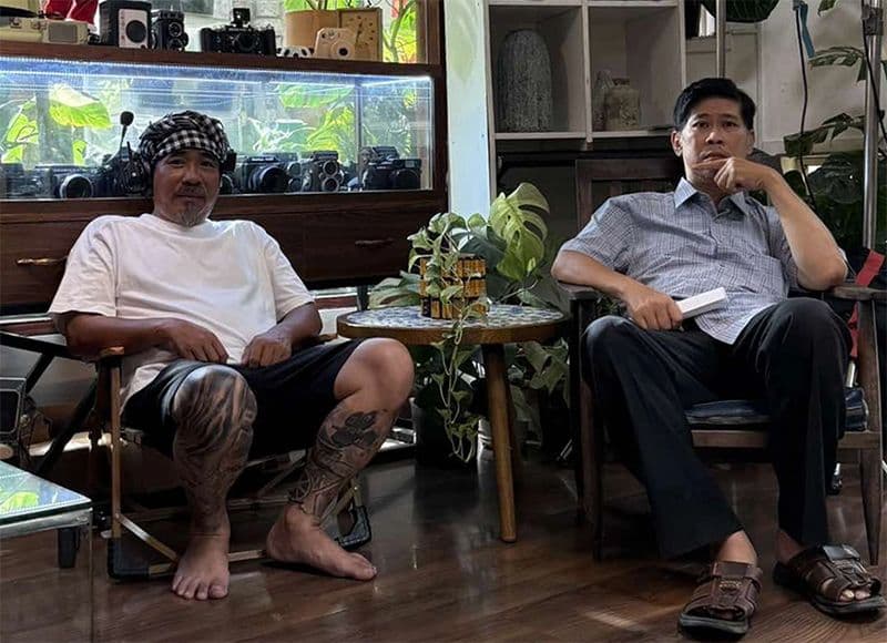 Nghệ sĩ Phước Sang ngồi cùng một đồng nghiệp trong không gian có nhiều máy ảnh và cây xanh, thể hiện sự trở lại phim trường và triết lý sống của anh.
