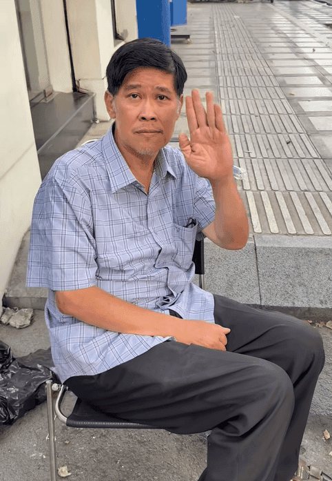 Nghệ sĩ Phước Sang ngồi trên ghế, giơ tay chào, thể hiện ý chí kiên cường sau biến cố sức khỏe và sự trở lại sân khấu.