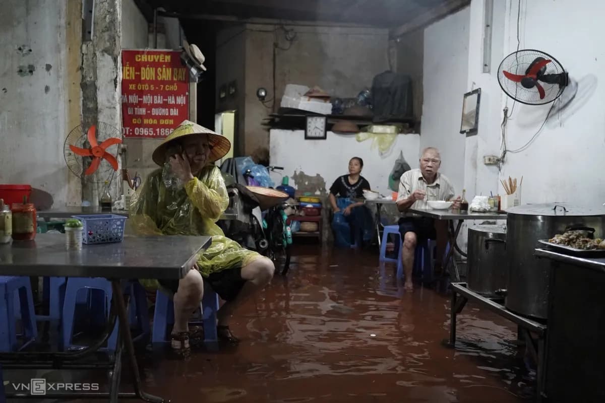 Người dân Hà Nội đội nón, mặc áo mưa ngồi ăn sáng trên vỉa hè cao ráo giữa dòng nước ngập.