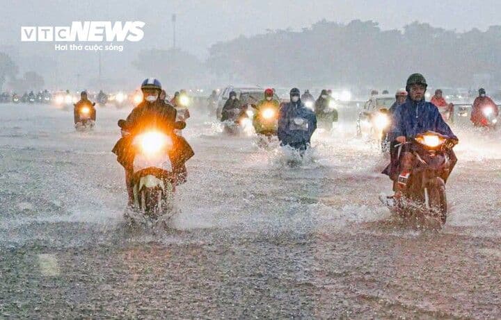 Người dân Hà Nội đi xe máy trong cơn mưa lớn trên đường ngập nước, thể hiện sự thích nghi với thời tiết khắc nghiệt.