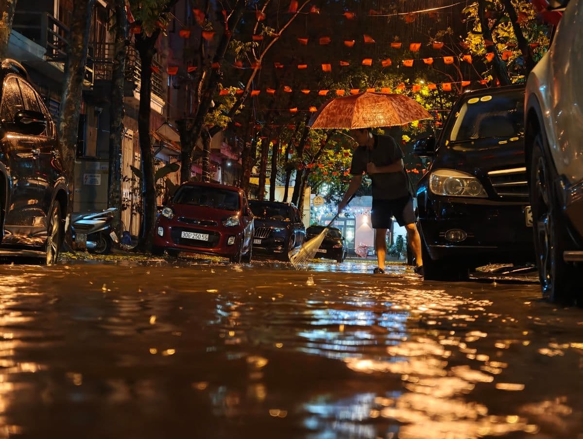 Người dân đội mưa dùng chổi khơi thông nắp cống trên đường phố ngập lụt ở Hà Nội.