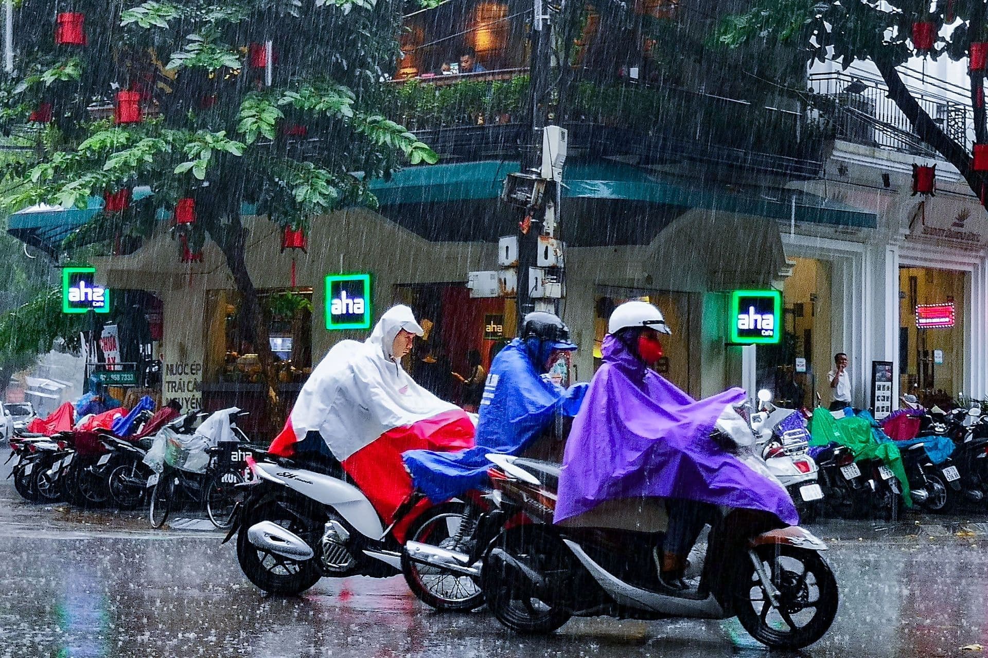 Người dân đi xe máy mặc áo mưa trên đường phố trong cơn mưa, thể hiện sự thích nghi hàng ngày với điều kiện thời tiết.