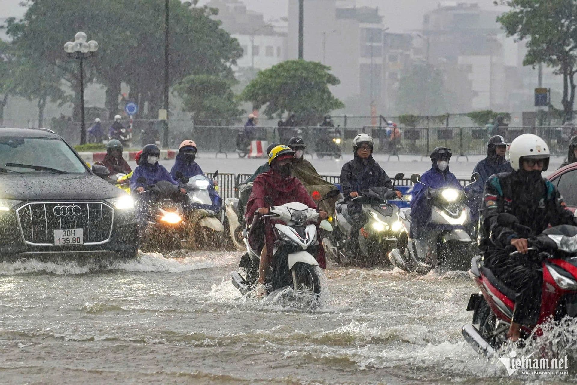 Người dân đi xe máy và ô tô qua đường phố ngập nước do mưa lớn ở Hà Nội, thể hiện tác động của thời tiết đến nhịp sống đô thị.
