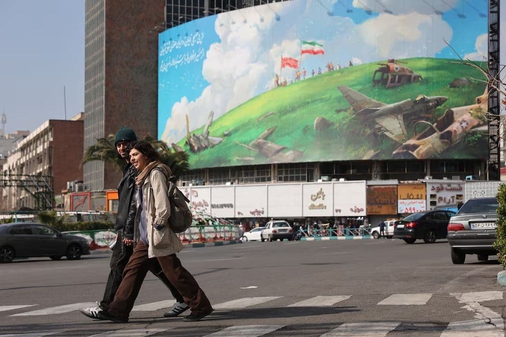 Người dân Iran đi bộ trên đường phố với biển quảng cáo lớn về máy bay quân sự bị phá hủy và cờ Iran, tượng trưng cho căng thẳng Mỹ-Iran.