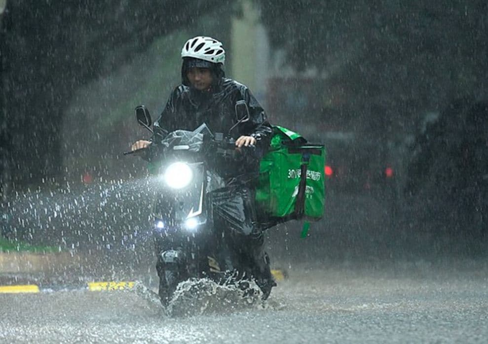 Người giao hàng đi xe máy trong mưa lớn, nước bắn tung tóe trên đường ngập lụt, thể hiện sự khẩn cấp và bình thường mới của thiên tai.
