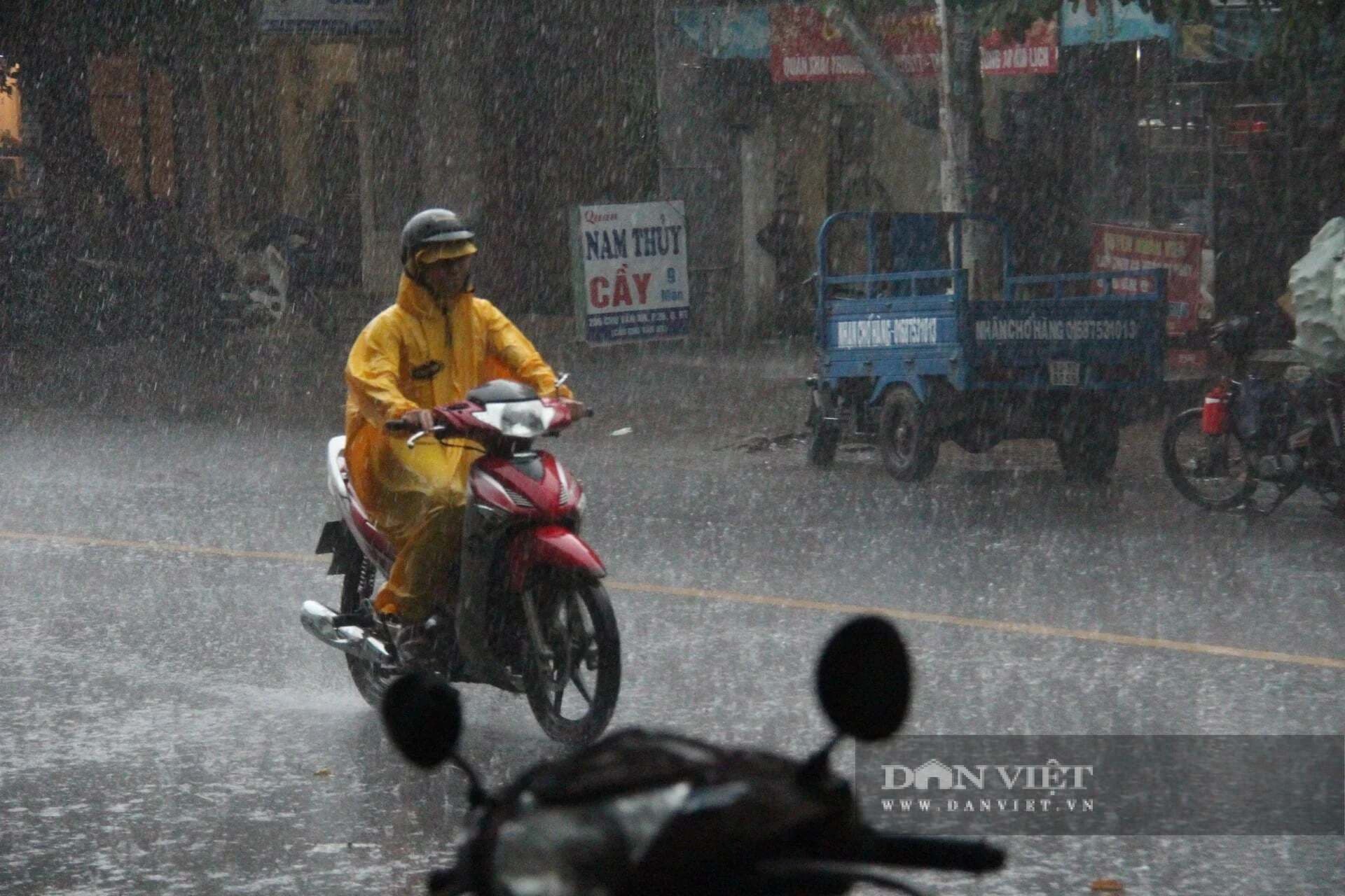 Người đi xe máy mặc áo mưa di chuyển trên đường phố Hà Nội trong cơn mưa lớn, thể hiện sự bất định của thời tiết.