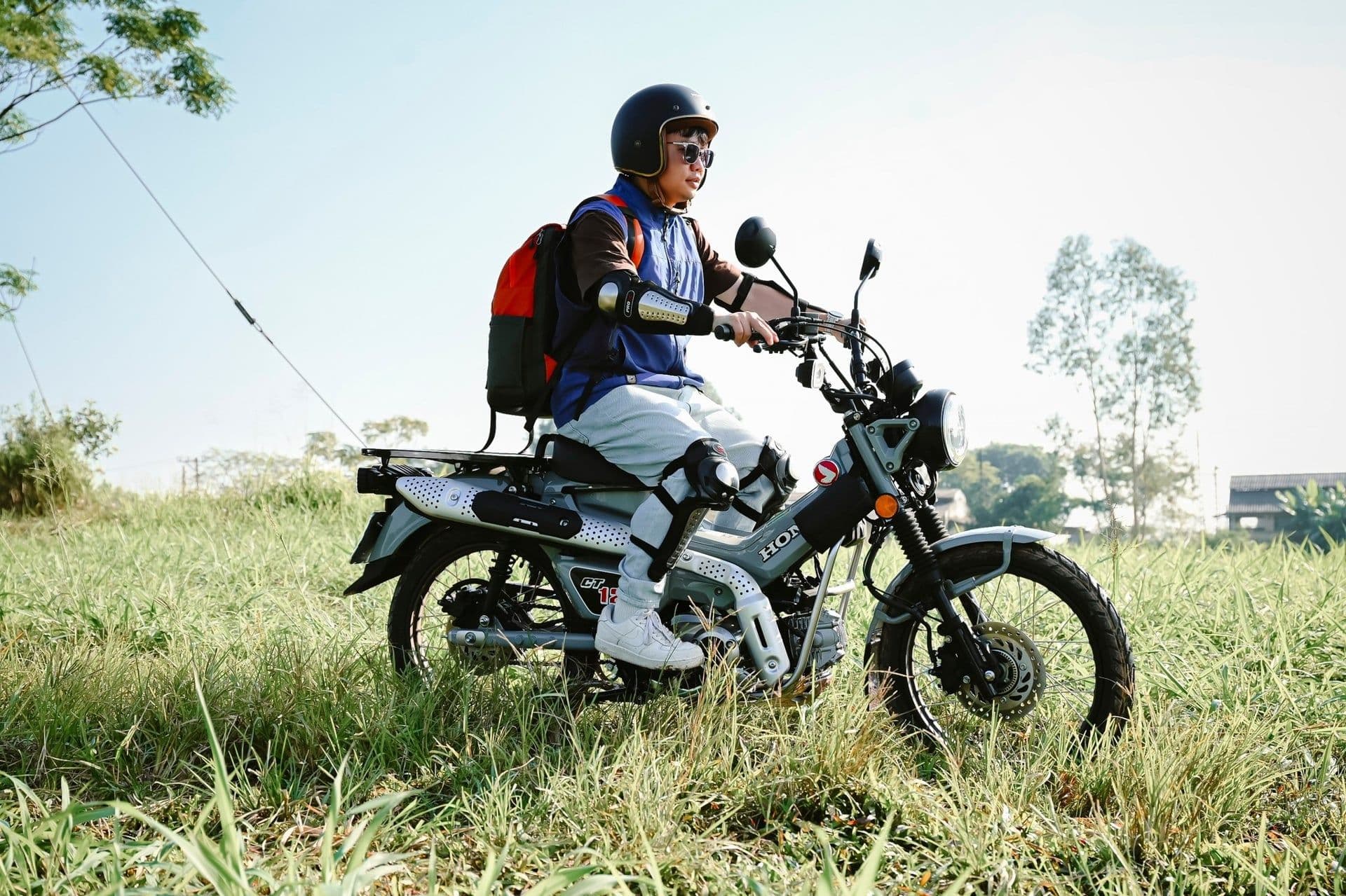 Người lái trên chiếc Honda CT125 màu xám bạc giữa cánh đồng, thể hiện tinh thần phiêu lưu và di sản của xe